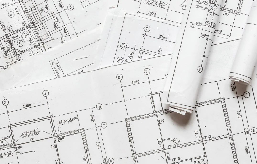 Desktop_About_Us_Legacy Several architectural blueprints with technical drawings, measurements, and floor plans are spread out, with some sheets rolled up on top.