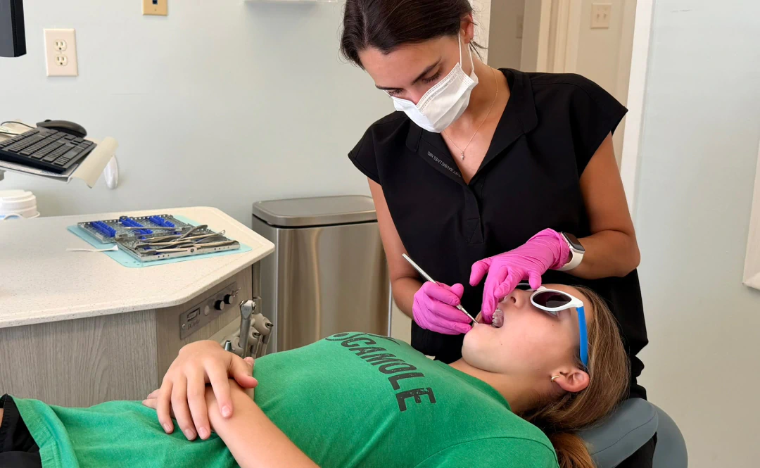 Desktop_About_Us_Rooted_In A dentist wearing gloves and a mask examines a patients teeth. The patient, lying back in the dental chair, wears sunglasses and a green shirt. Dental tools are on a tray nearby.