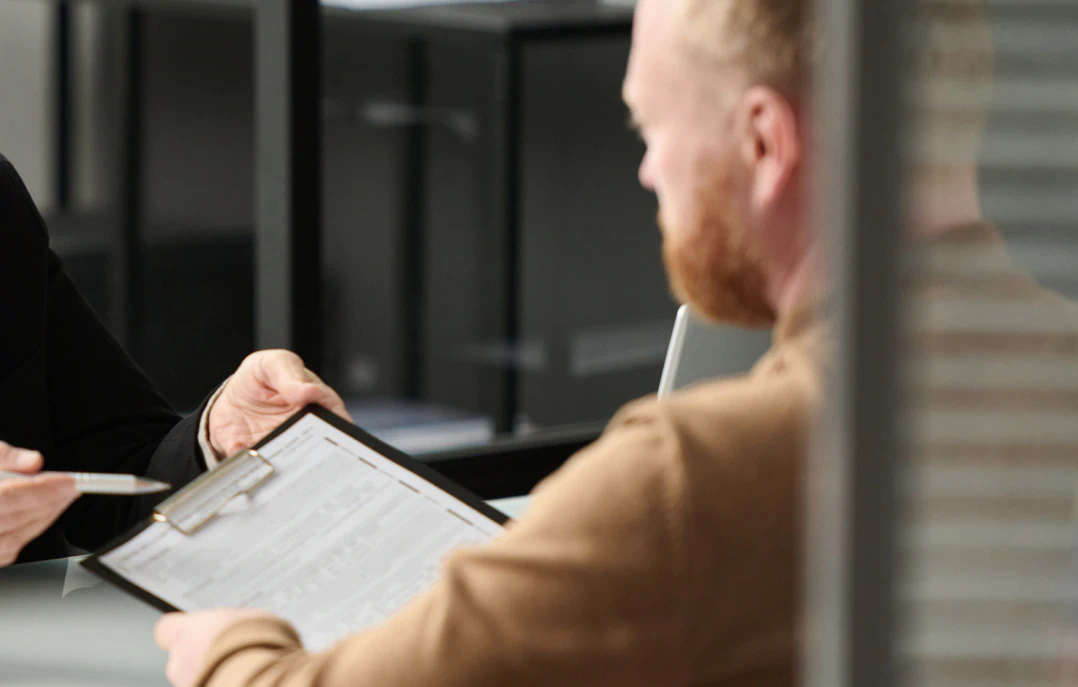 Desktop_About_Us_Stewardship A person hands a clipboard with a form to another person, who is seated at a desk. The background shows office furniture and blurred partitions.