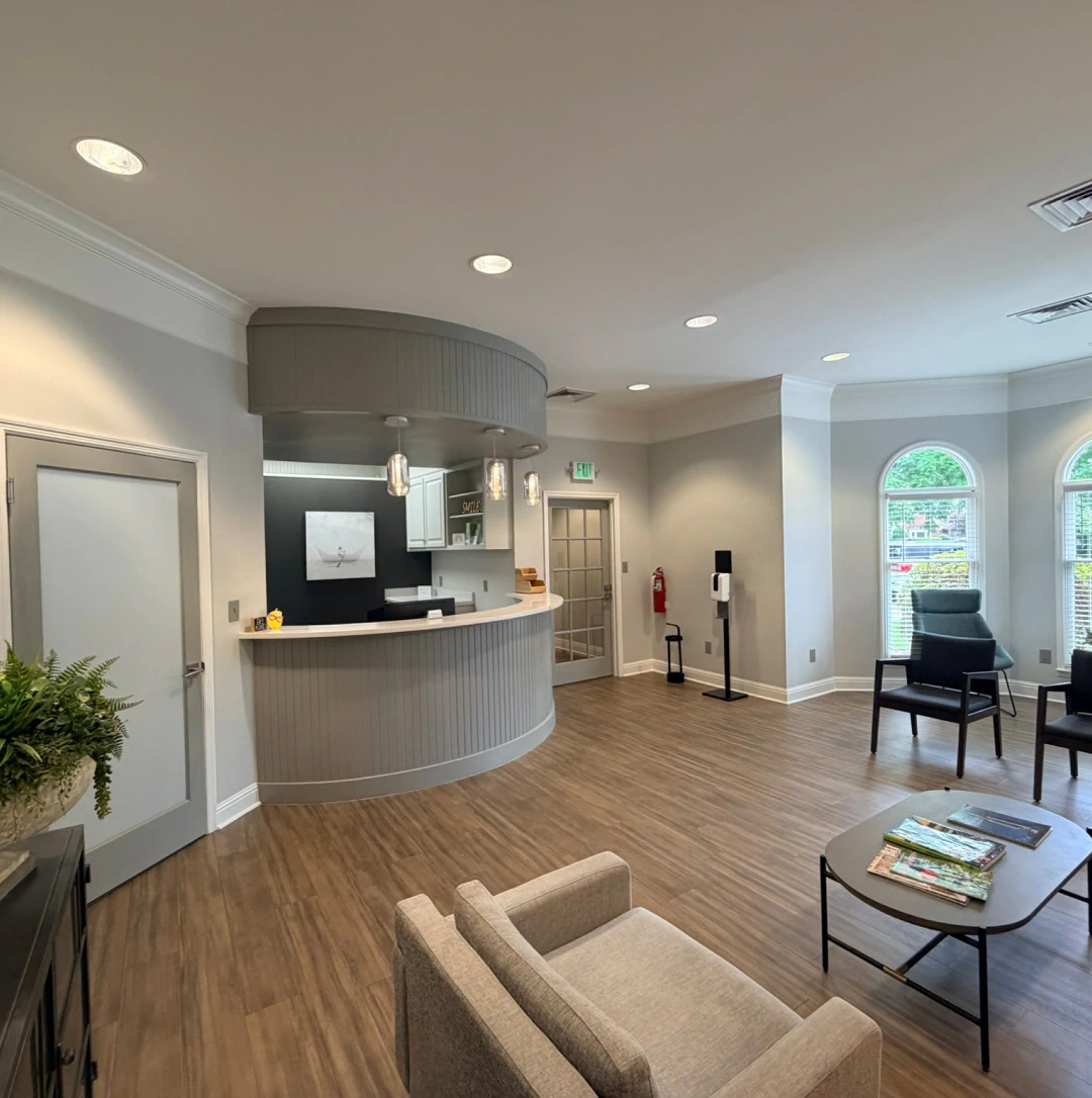 Desktop_About_Us_Strategic_Advocacy A modern office lobby with wood flooring, a curved reception desk, chairs, magazines on tables, large windows, and neutral-colored walls, creating a welcoming atmosphere.