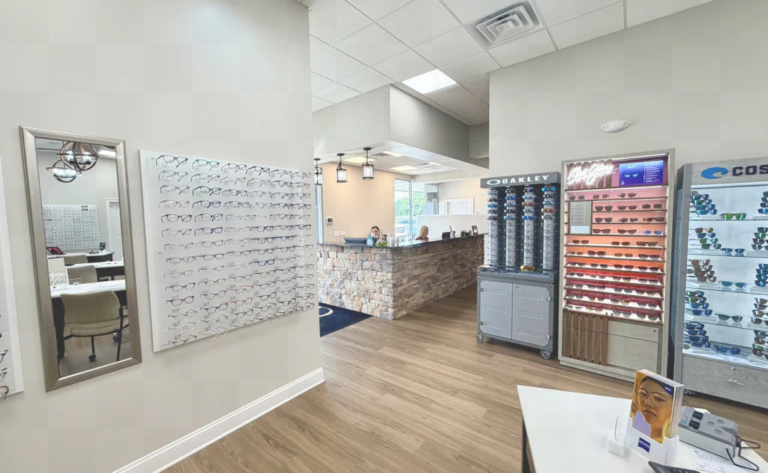 Desktop_Contact_Us_Lets_Make An eyeglass store interior with display racks of glasses, a counter with stone detailing, a staff member behind the counter, and light wood flooring. Bright lighting and modern decor are visible throughout the space.
