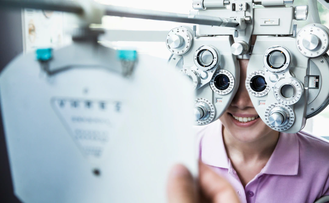Desktop_Meet_the_Doctor_Committed_to A person in a pink shirt smiles while having their vision tested with a phoropter. An eye chart is visible in the foreground, slightly out of focus.