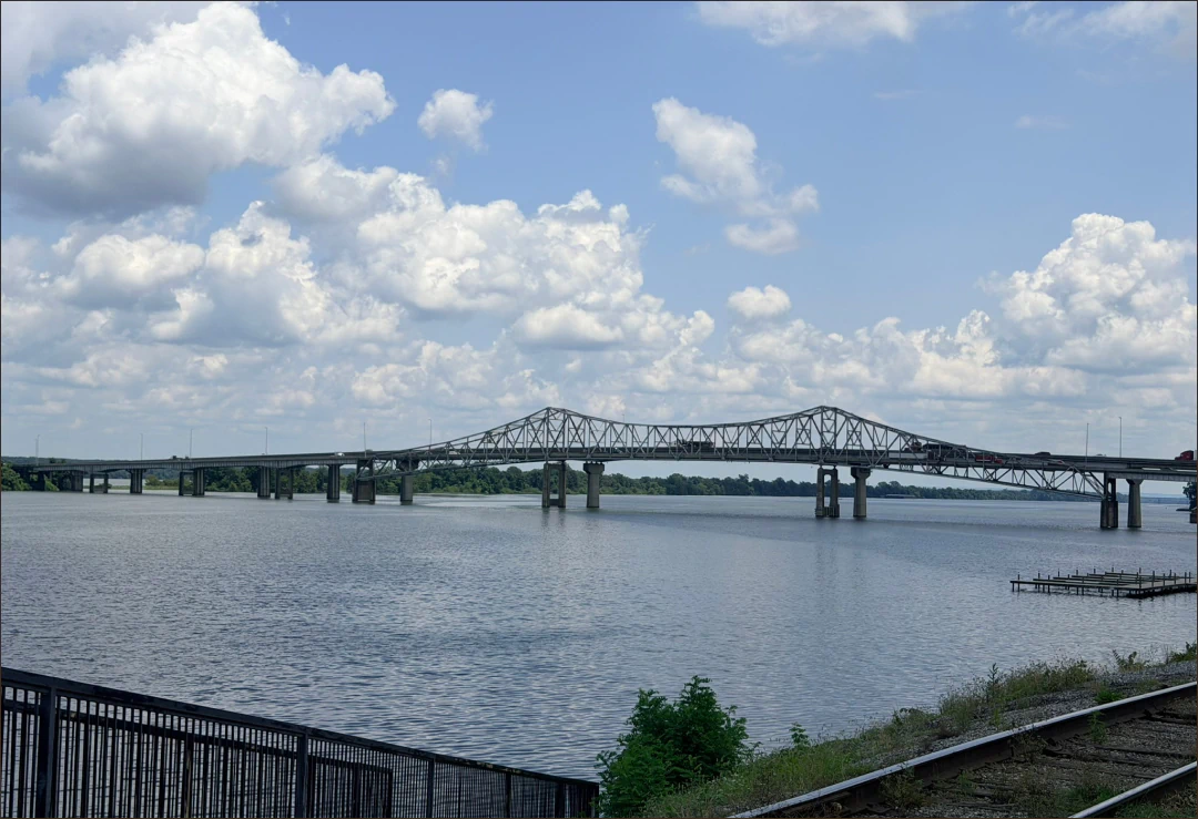 Desktop_Meet_the_Doctor_Personal_Life A steel truss bridge spans a wide river under a partly cloudy sky, with a railing and railway tracks in the foreground and green trees in the distance.