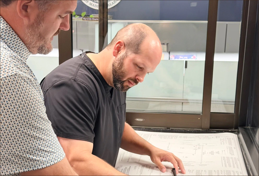 Desktop_Meet_the_Doctor_Why_He Two men stand by a window reviewing blueprints or architectural plans spread out on a table, with one man pointing at the documents and the other observing.